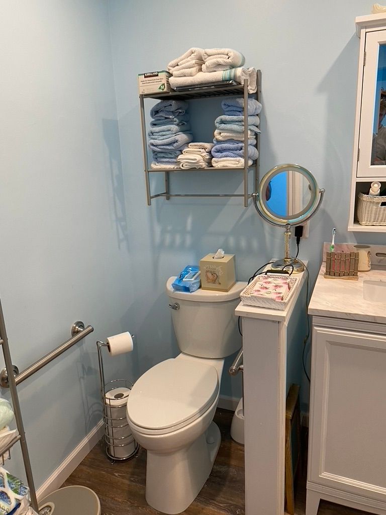 Small bathroom with toilet, shelves of towels, and a vanity. Light blue walls and wood-look floor.