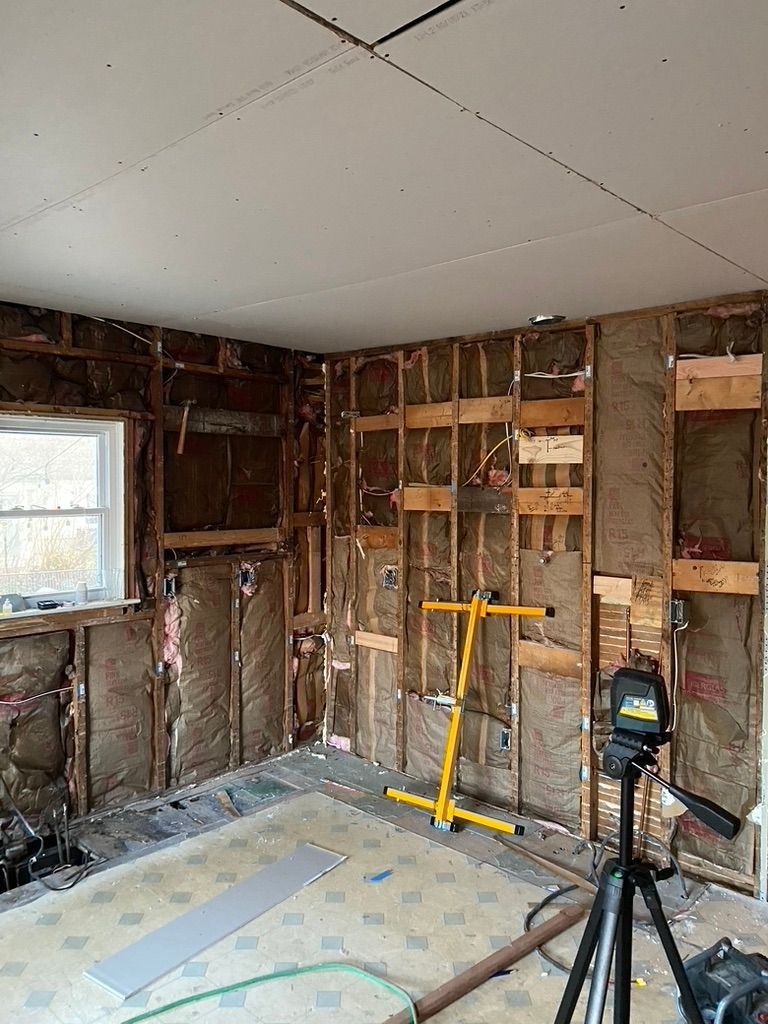 Room under renovation with exposed wooden framing and insulation. Yellow work light and a laser level on a tripod are visible.