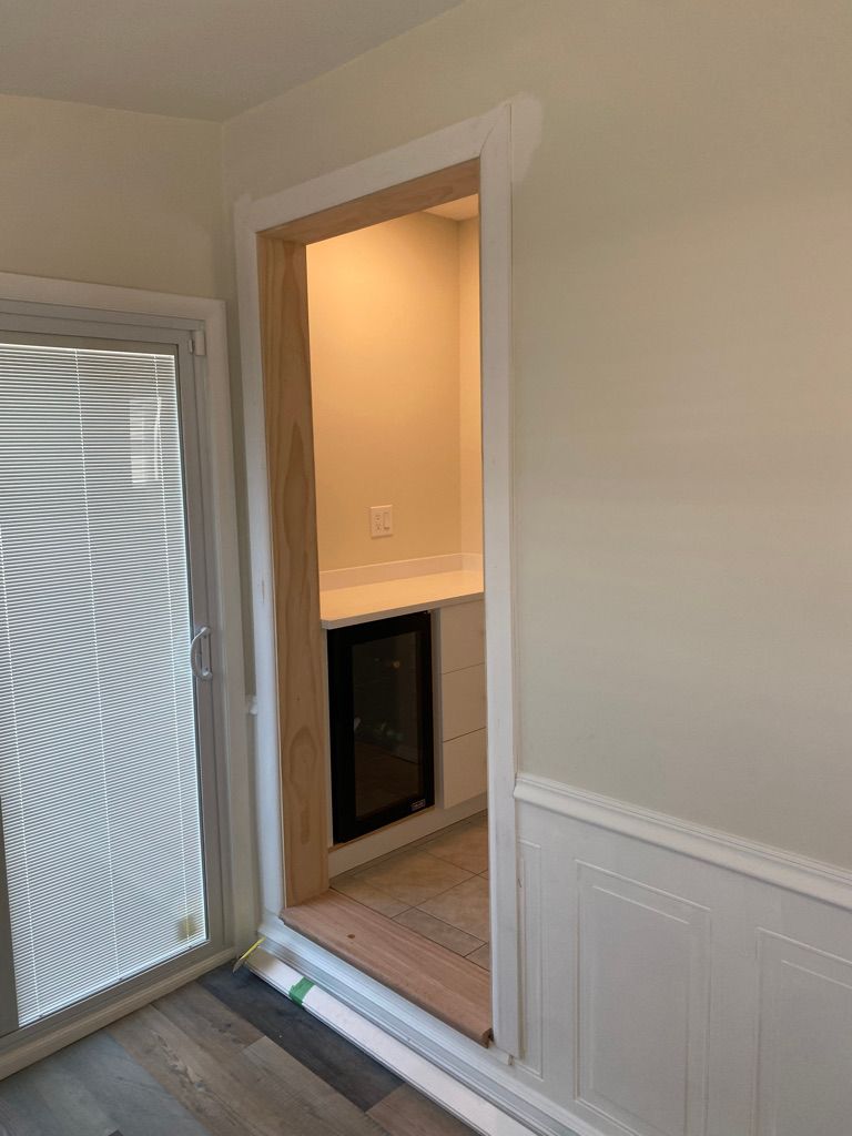 View into a small room, with a wine fridge, from a doorway. White trim, beige walls, gray floor.