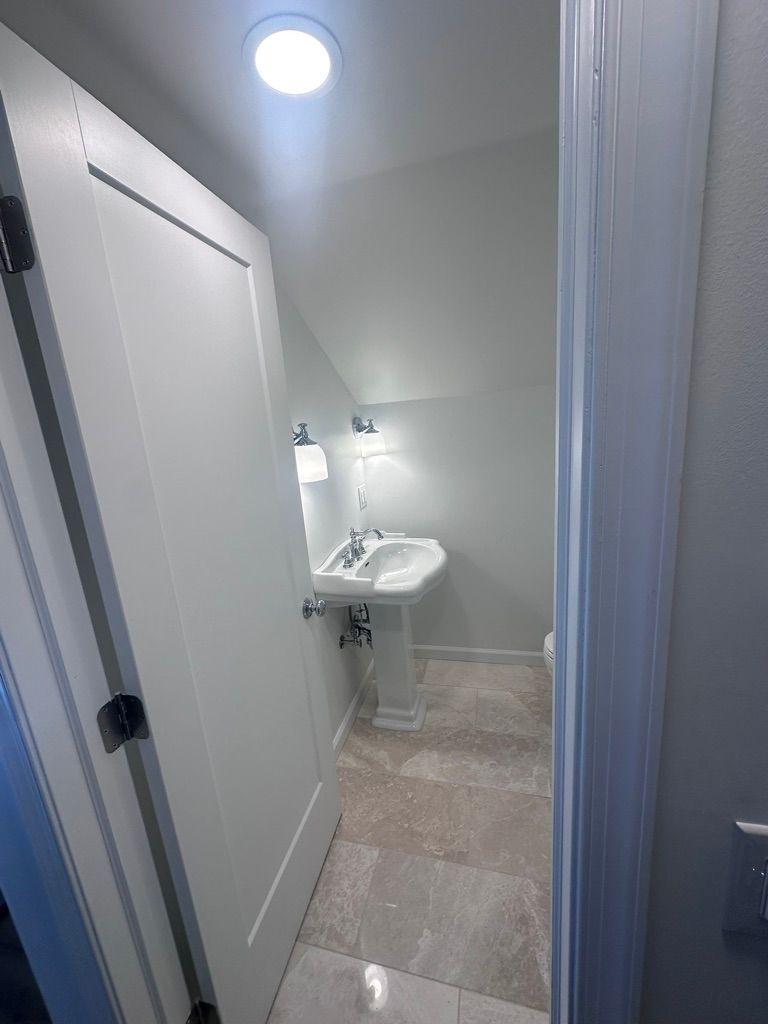 Narrow bathroom with pedestal sink, light-colored walls, open door, and tile floor.