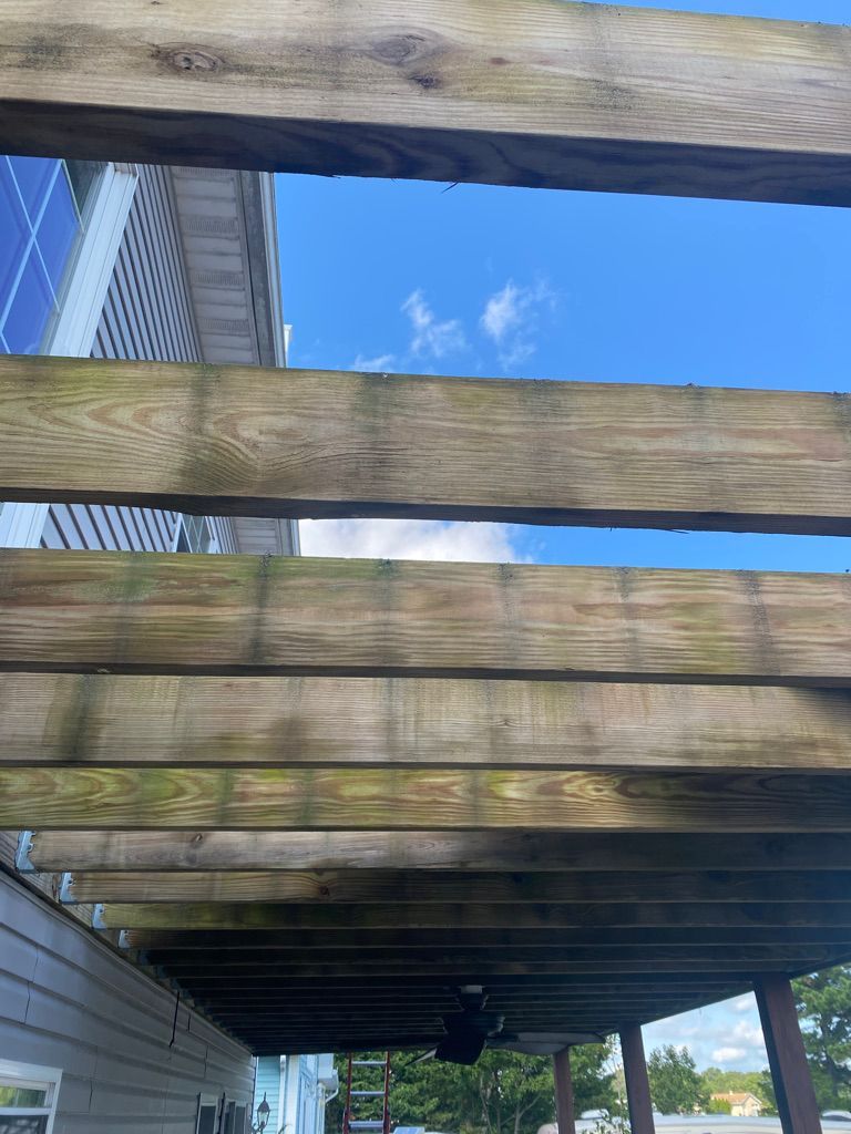 Wooden beams of a pergola against a blue sky with clouds. Below, a section of a house is visible.