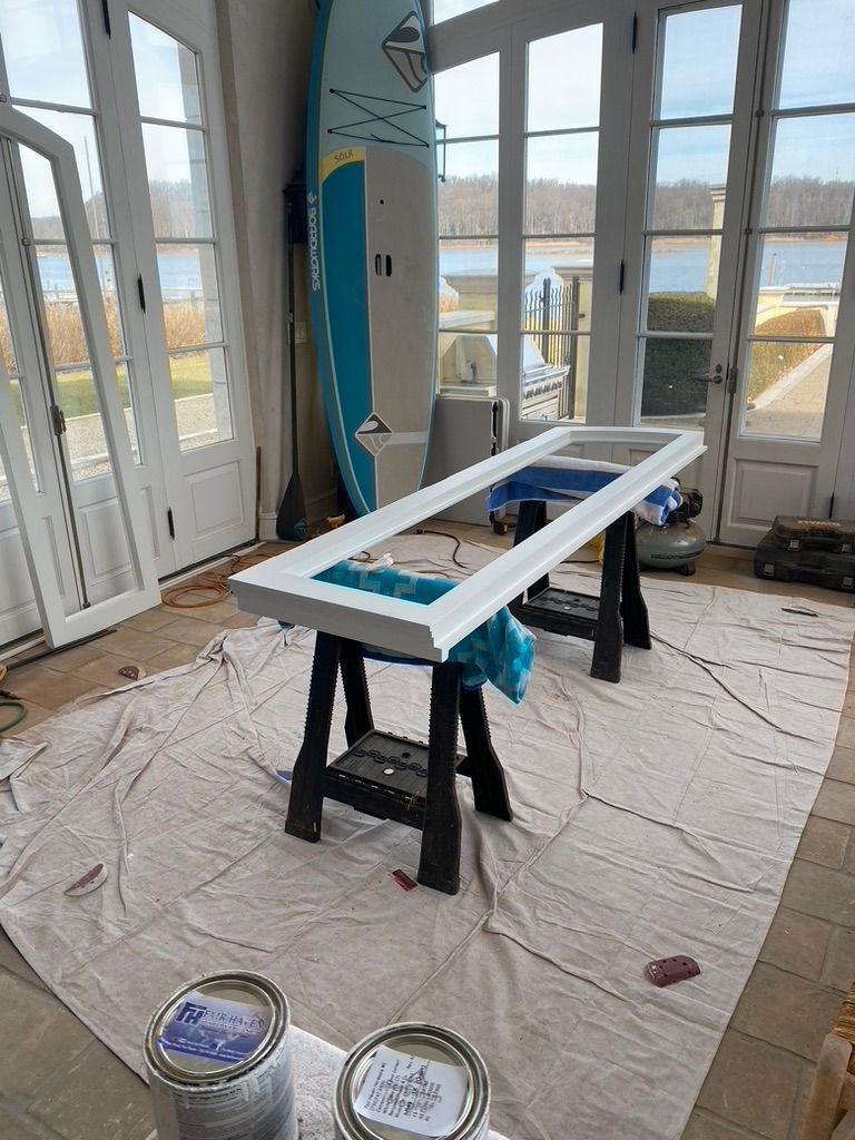 White-painted door frame on sawhorses in sunroom with water view, paint cans, and paddleboard.