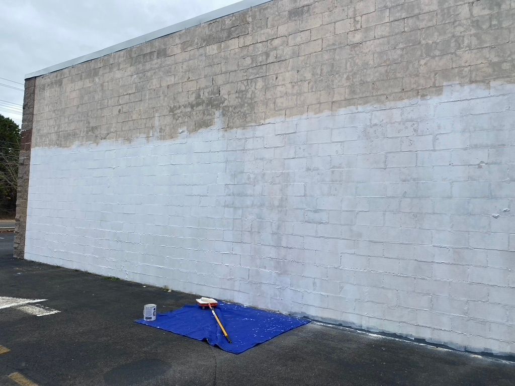 A brick wall partially painted white, with paint supplies on a blue tarp in front of it.