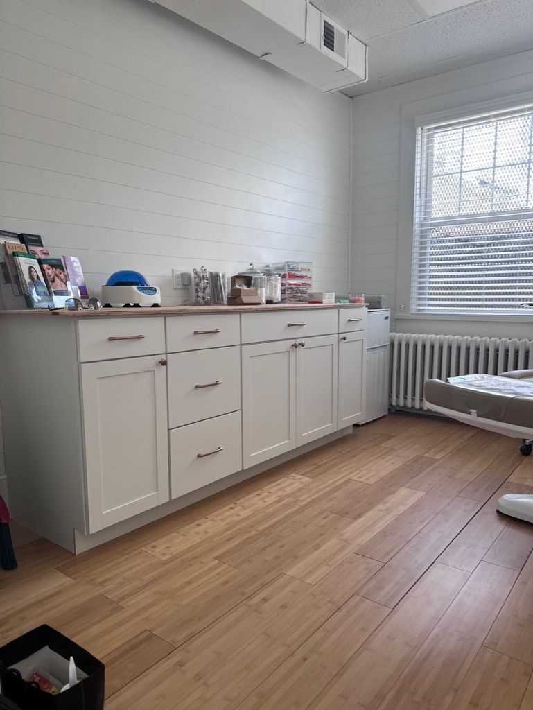 White cabinets with wood countertop against a white wall; wooden floor; window with blinds.