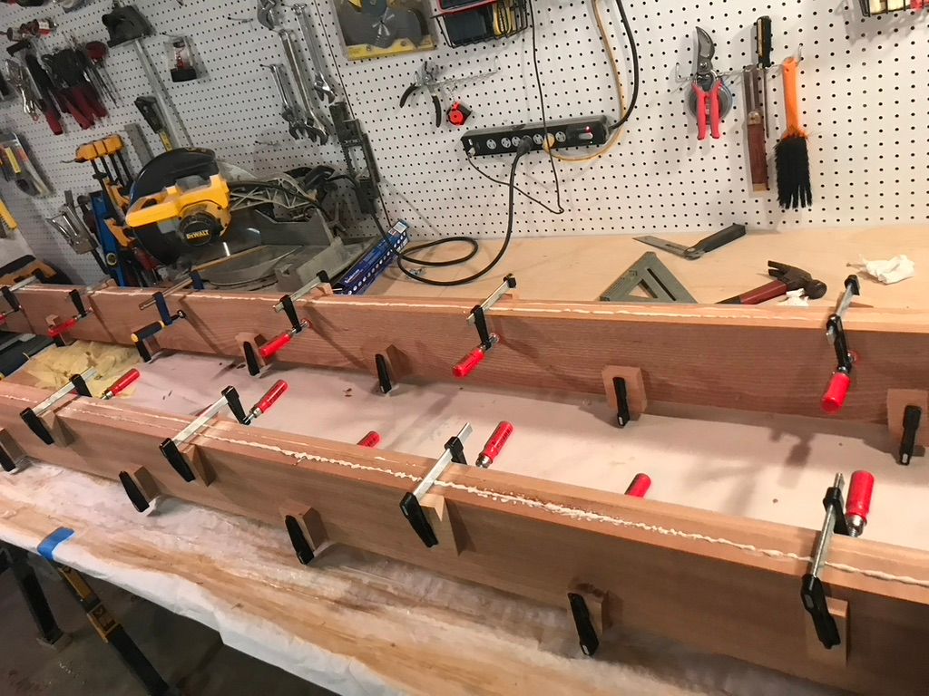 Wood pieces clamped together on a workbench, with glue visible. Workshop tools hang in background.