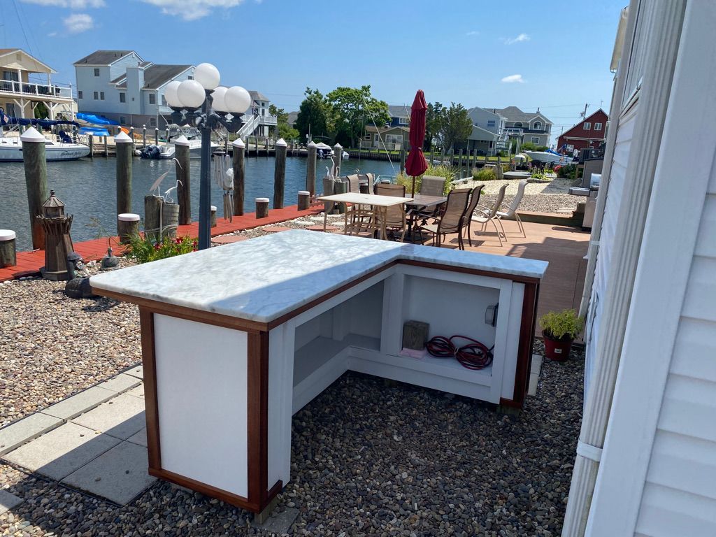 L-shaped outdoor bar with white countertop and cabinets, wooden trim, set on a stone patio overlooking a waterway.