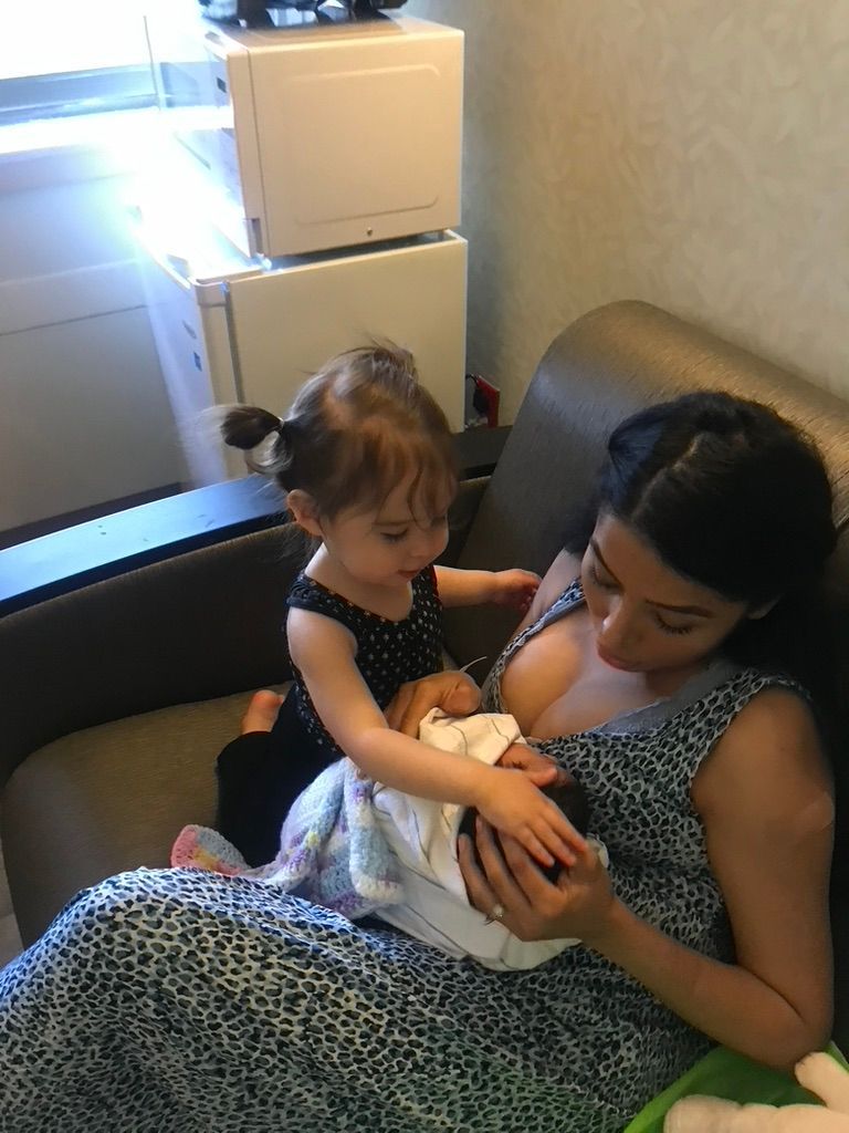 Woman breastfeeding a baby on a couch. A toddler watches, touching the baby's blanket. Light interior setting.