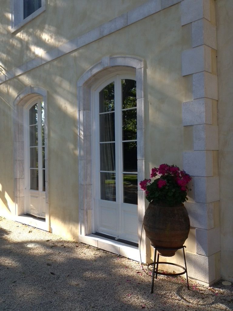 Exterior of a building with arched white door and window, stone trim, potted pink flowers, and sunlight.