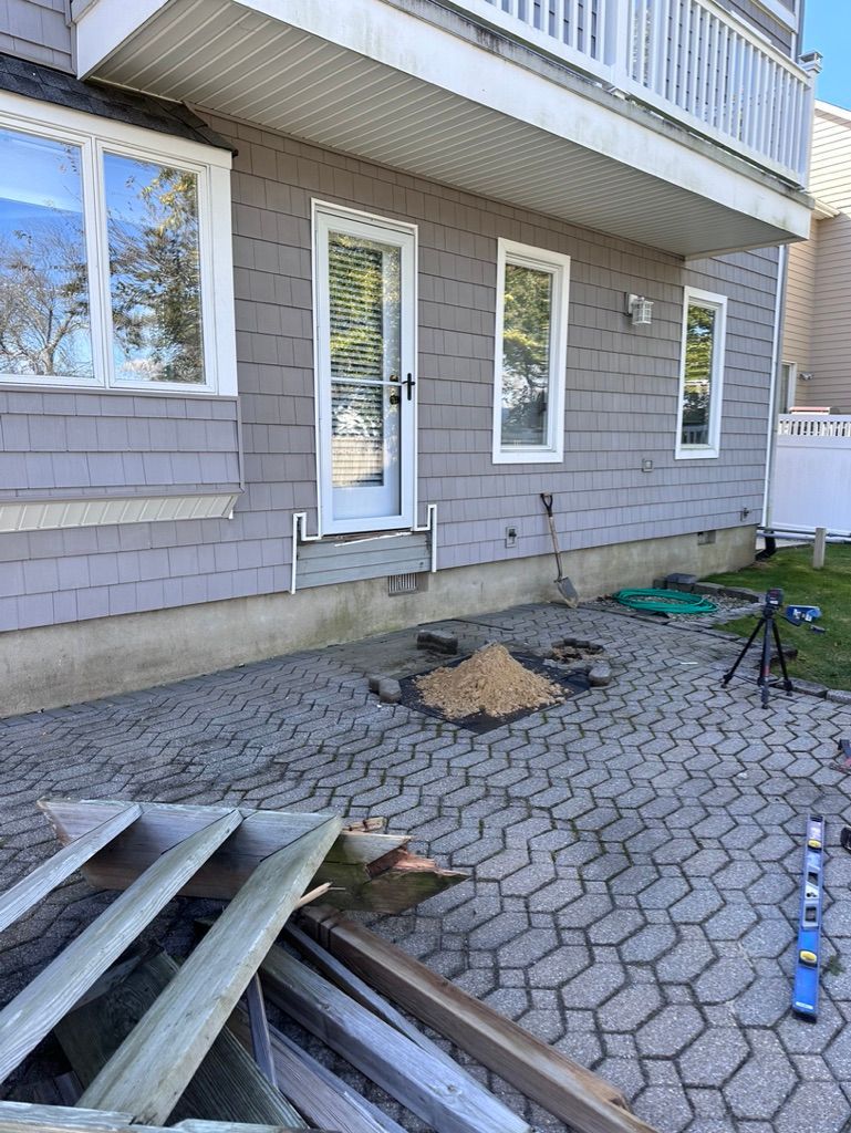 Exterior of a house with gray siding, white windows, and a paved patio; lumber and tools are nearby.