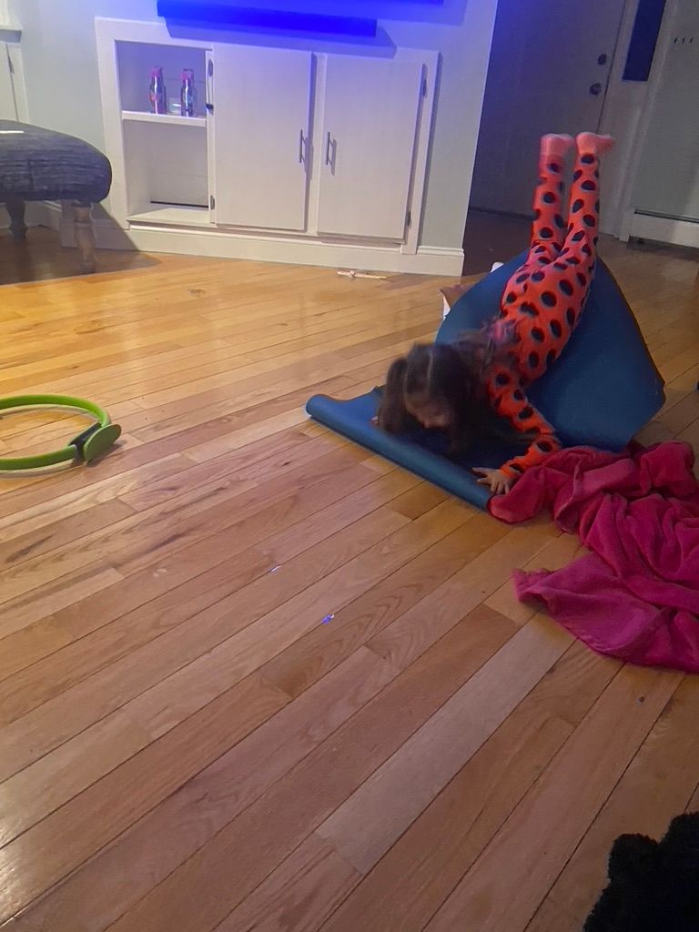 Child in orange and black leggings does a handstand on a yoga mat near a blue object; hardwood floor.