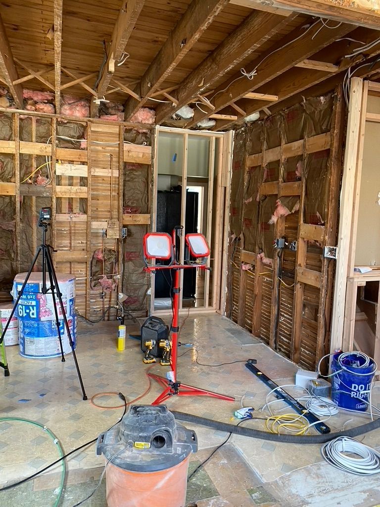 Interior construction site. Walls stripped down to studs. Work lights, tools, insulation visible.