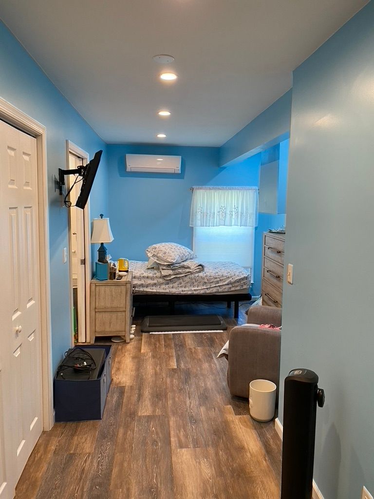Long hallway with light blue walls, leading to a bedroom with bed and dresser; wood flooring.