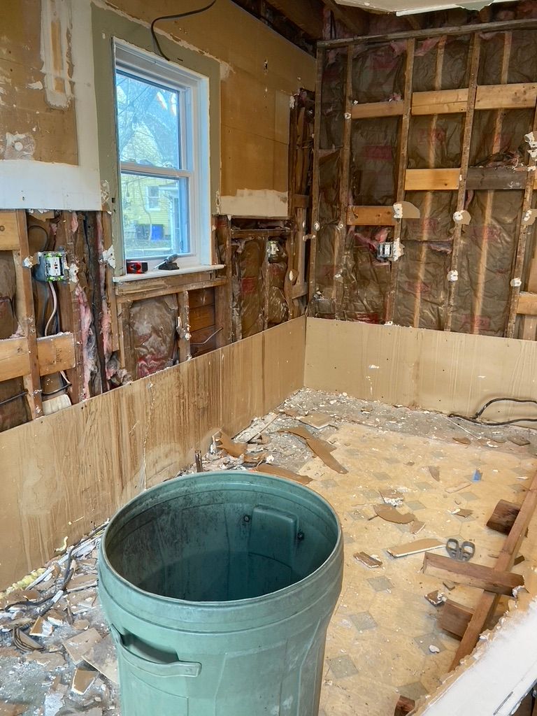 Room undergoing renovation: exposed studs, debris, green trash can, window, and torn walls.