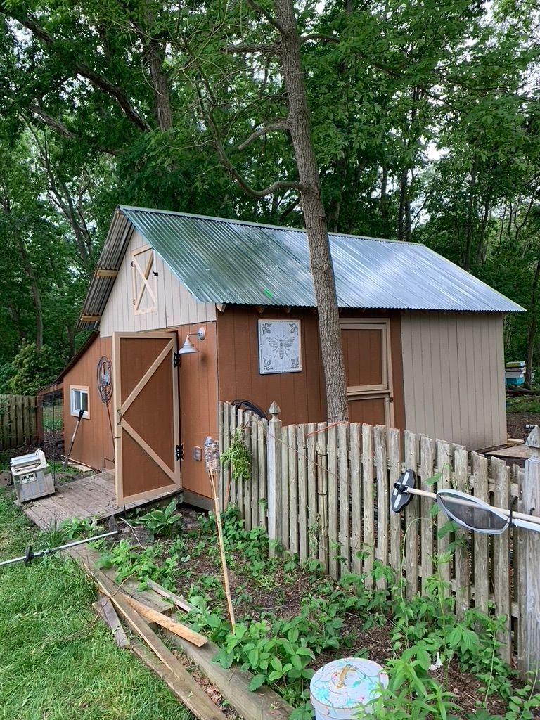 Small brown and tan shed with a corrugated metal roof, next to a wooden fence and surrounded by trees.