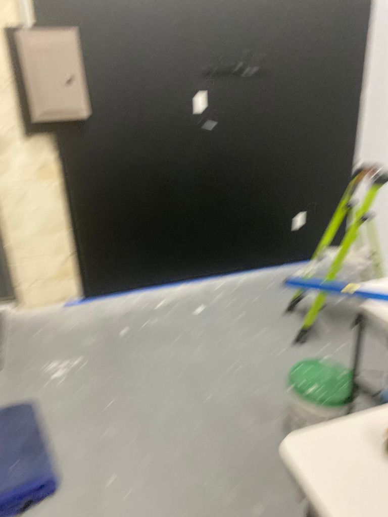 A room with a painted black wall, a step ladder, and a gray floor with paint flecks.