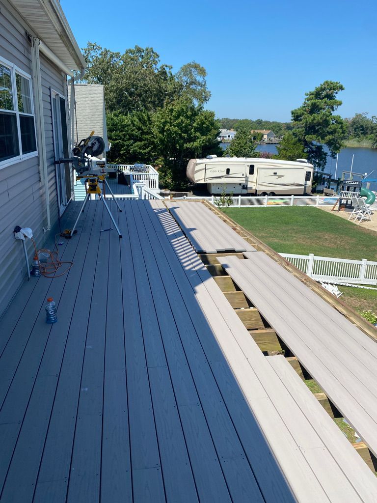 A deck with missing planks next to a house, with a water view and a parked RV.