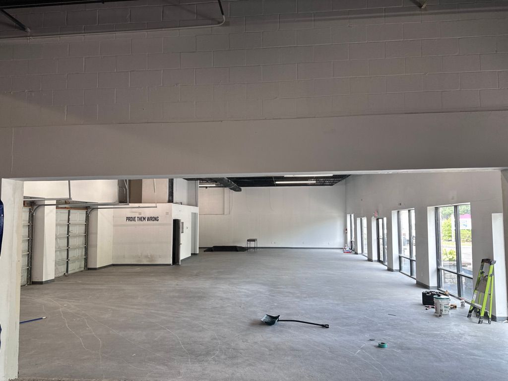 Empty commercial space with concrete floor, white walls, and large windows. Construction tools present.