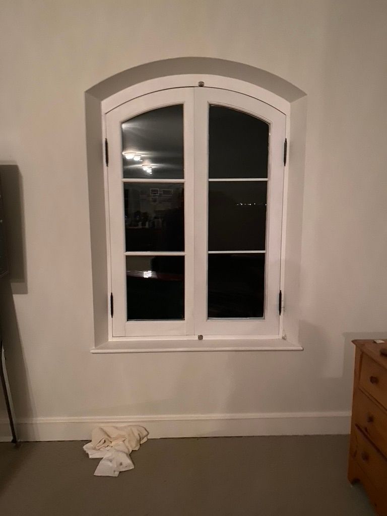White arched window in a recessed wall, reflecting nighttime lights.  White trim and a small drawer chest are visible.