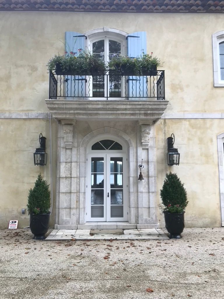 Exterior view of a cream-colored building with a balcony, double white doors, and lanterns. Two potted trees flank the door.