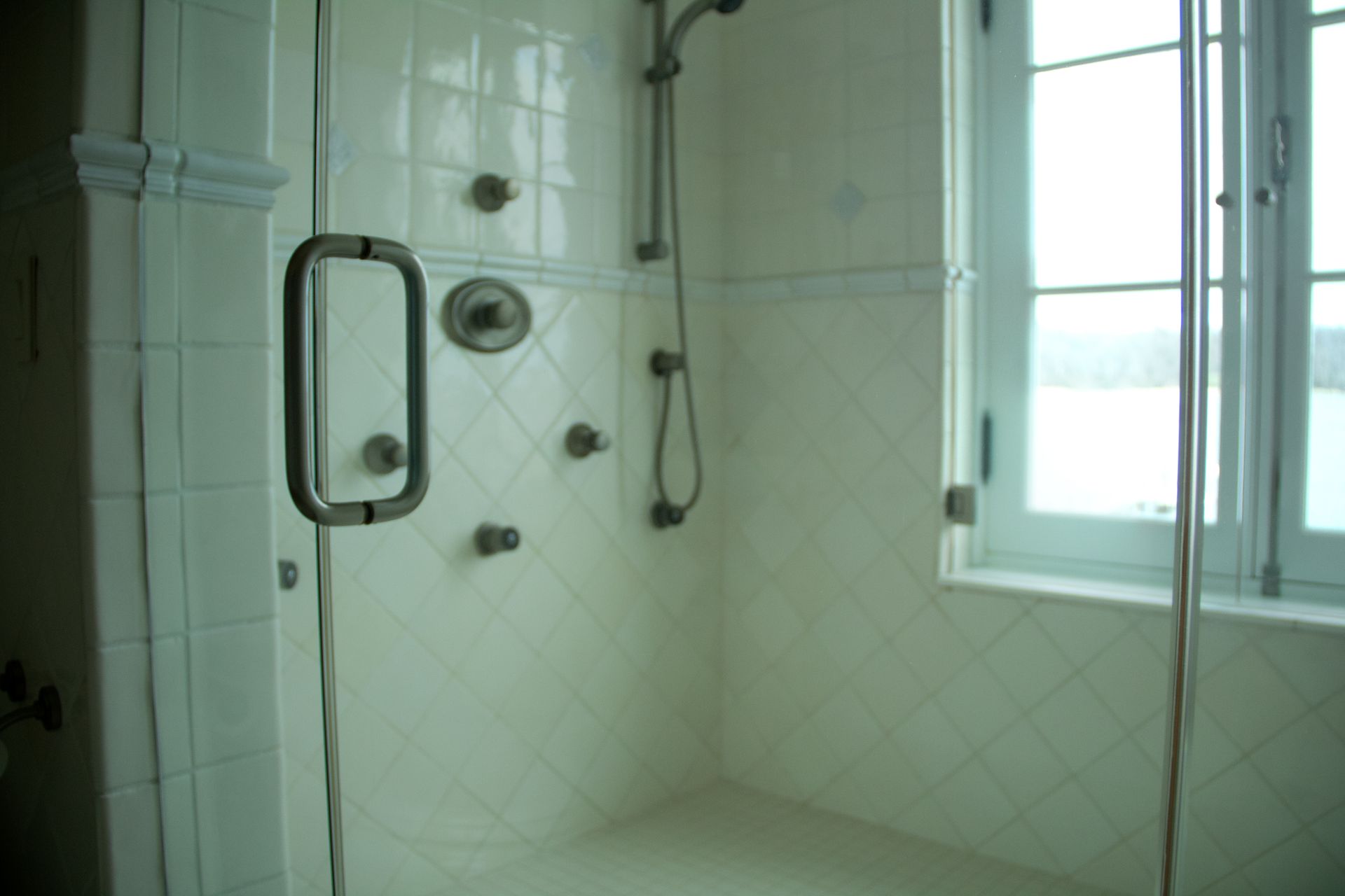 Glass-walled shower with white tiled walls, multiple shower heads, and window.