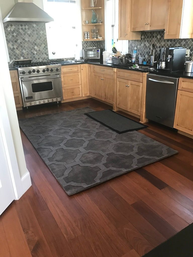 A kitchen with light wood cabinets, dark granite countertops, stainless steel appliances, and a gray patterned area rug.
