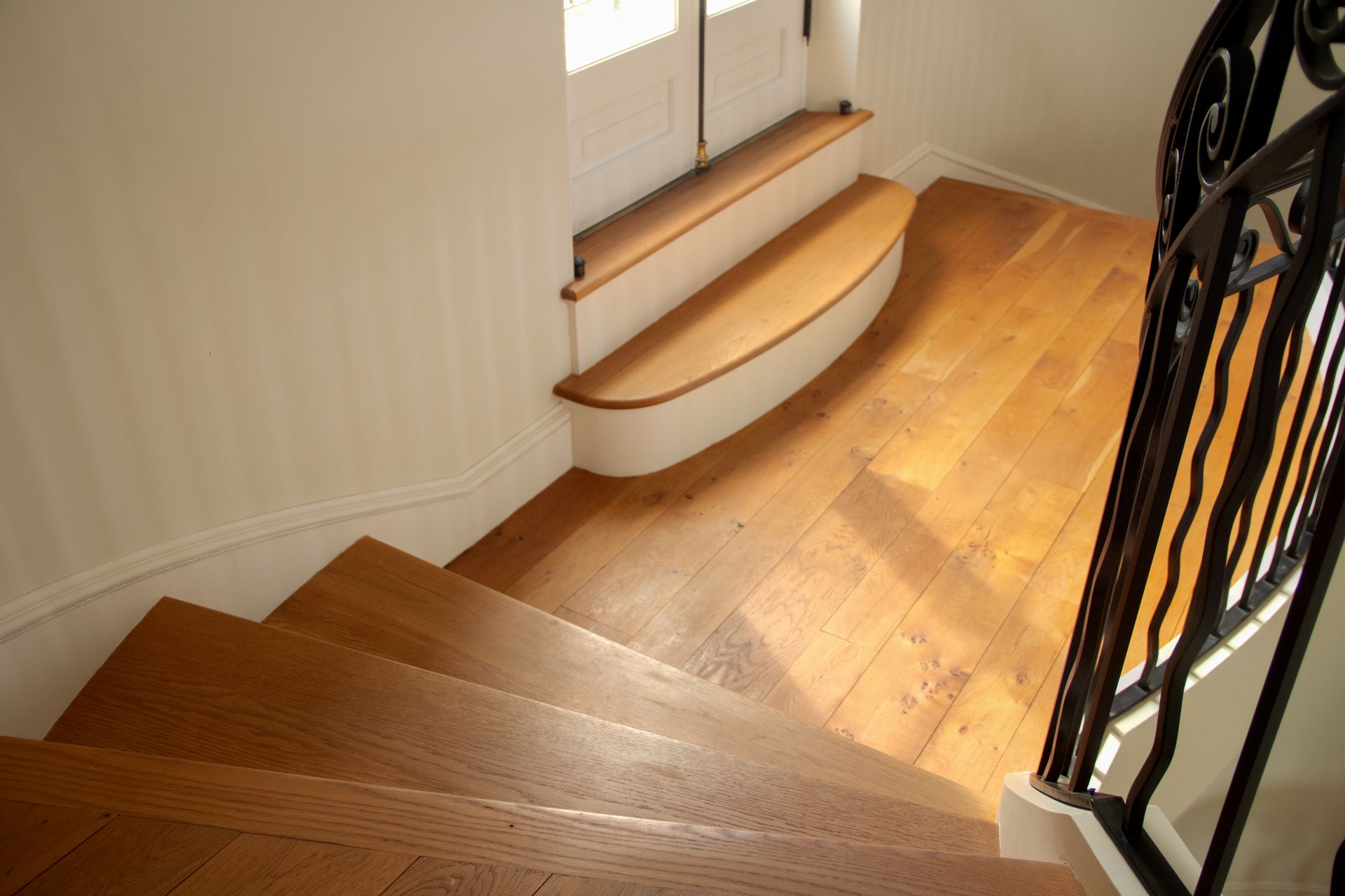 Wooden staircase with a curved landing, black railing, sunlight.