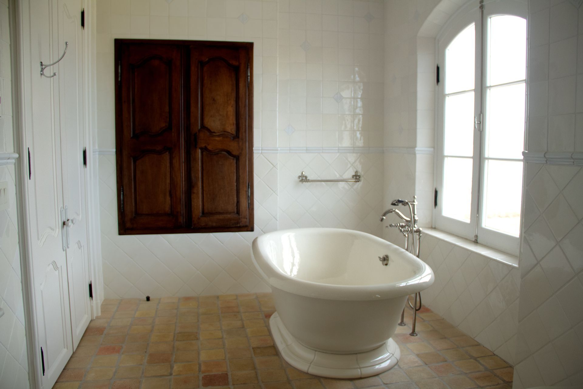 Bathroom with white walls, arched window, wooden shutters, and a freestanding white bathtub.