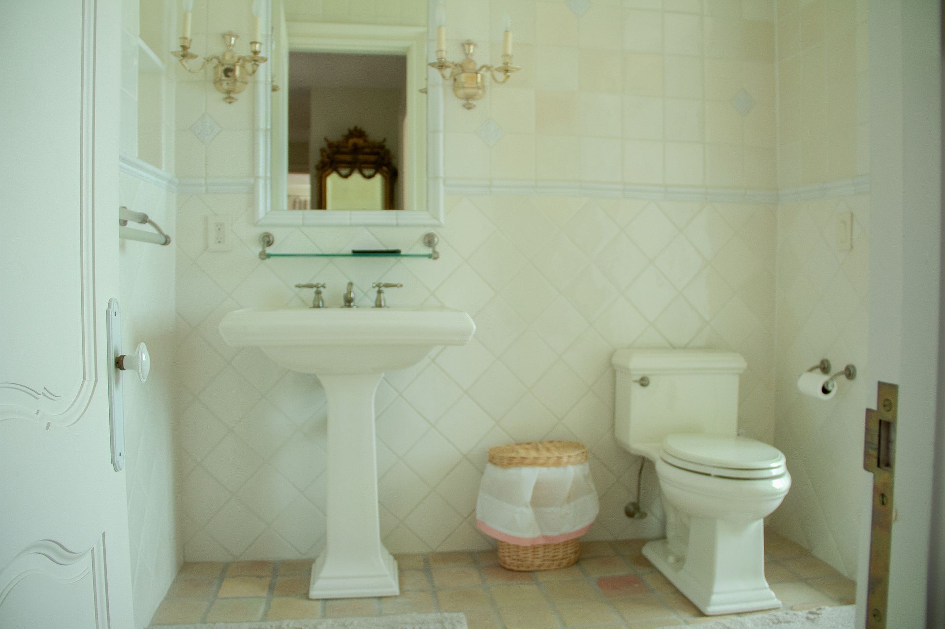 White bathroom with pedestal sink, toilet, mirror, and wicker trash can.