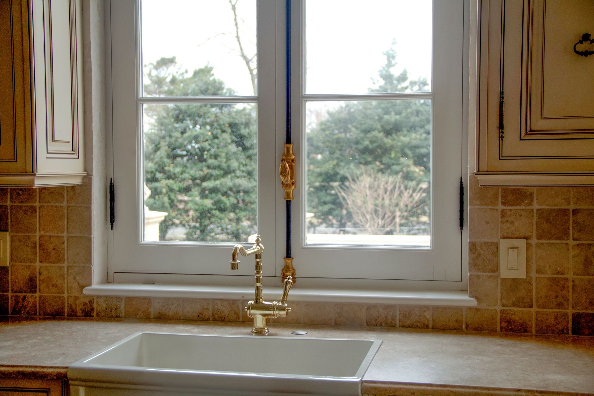 Kitchen sink under a window with trees outside. Gold faucet, white sink, beige tile and cabinets.