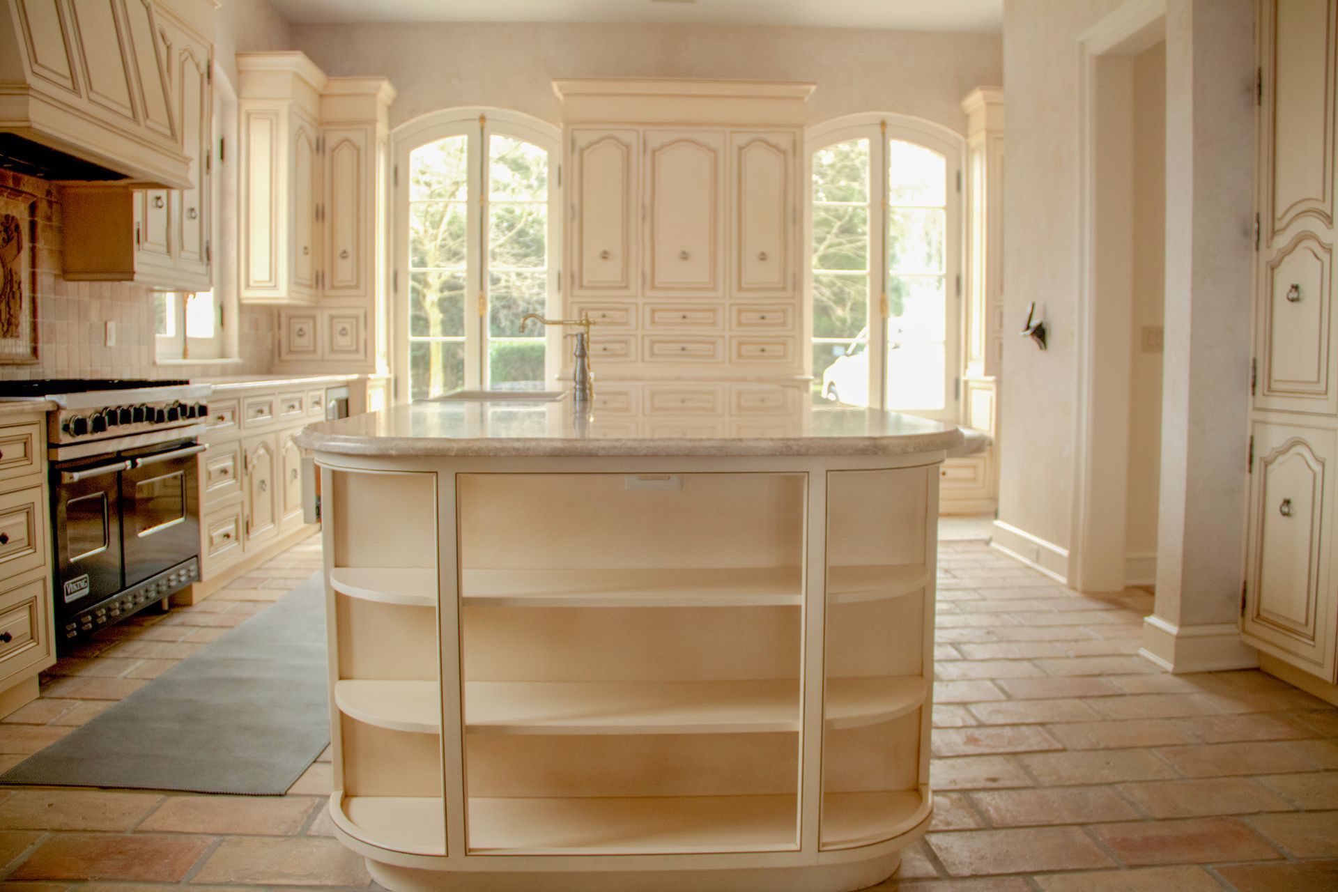 Cream-colored kitchen with an island, oven, and large windows. Stone floor and cabinets.