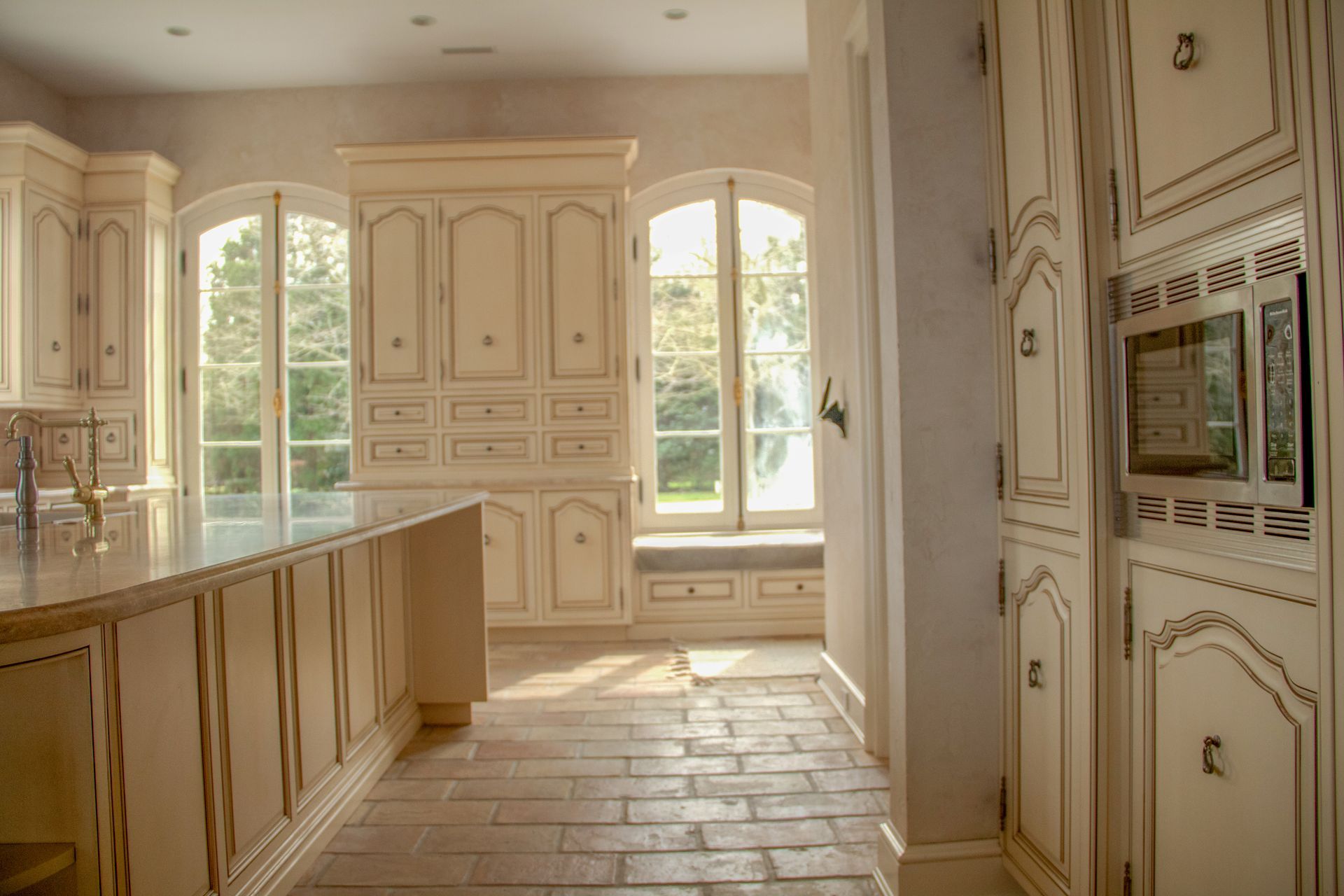 Cream-colored kitchen with arched windows, light shining on a brick floor. Built-in cabinets and appliances.
