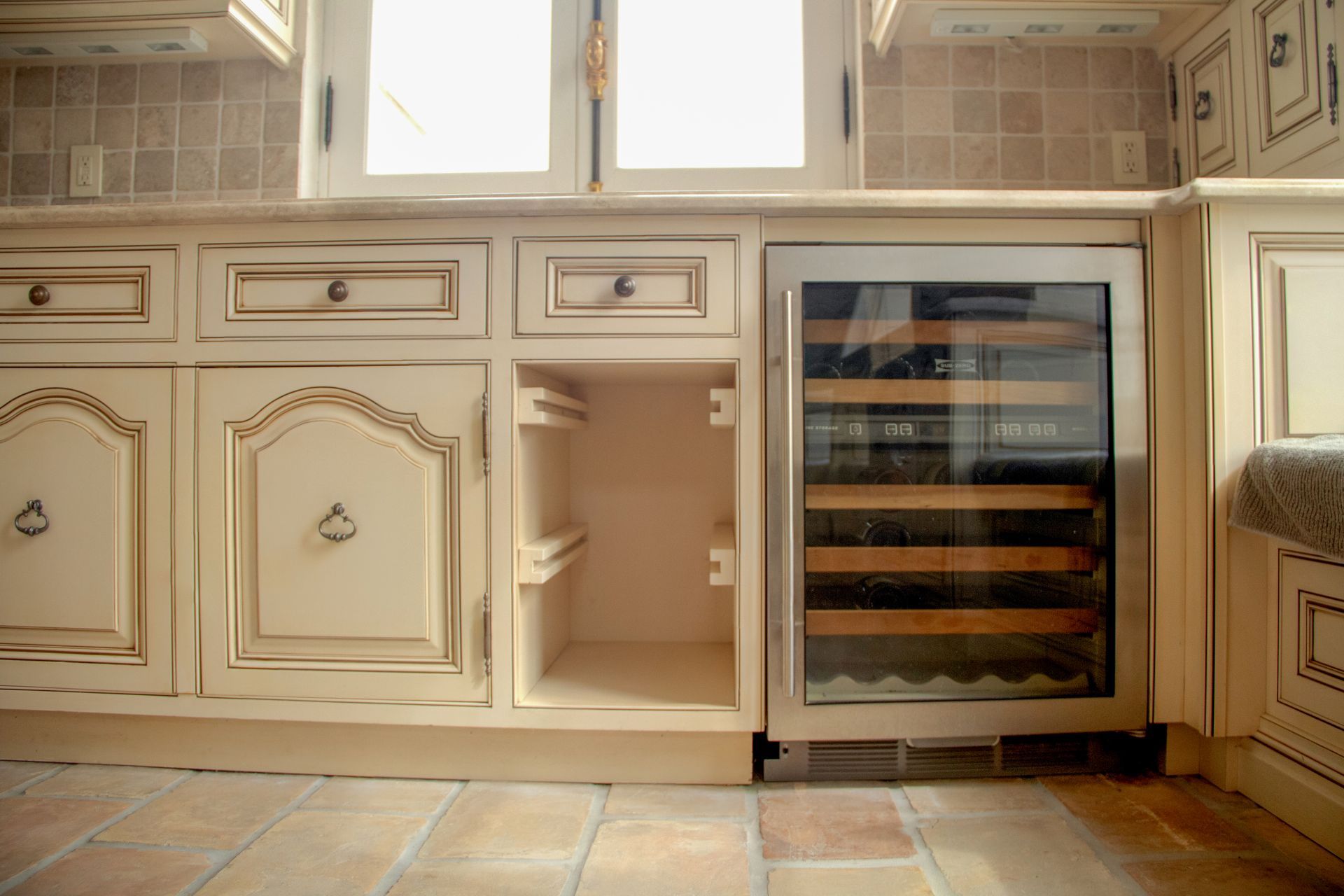 Cream-colored kitchen cabinets with a stainless steel wine cooler built-in. Beige tile backsplash and a brown tile floor.