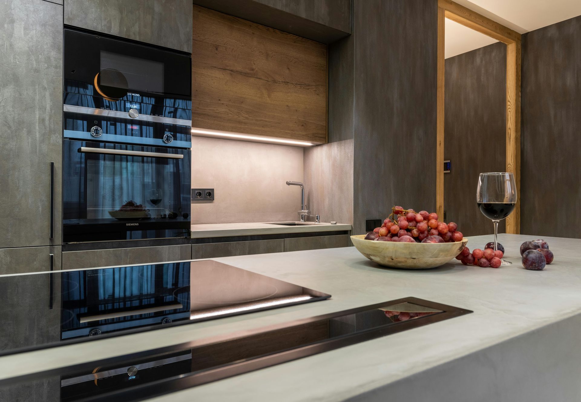 A modern kitchen island with a bowl of grapes and a glass of red wine, featuring a sleek, dark built-in oven and backsplash.