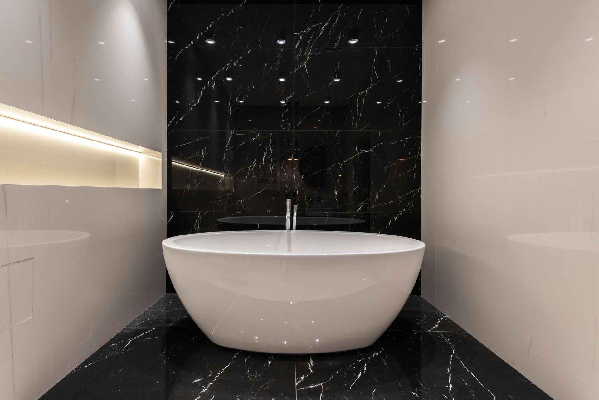 A modern white oval bathtub centered against a black marble wall in a bathroom with glossy white side walls and warm lighting.