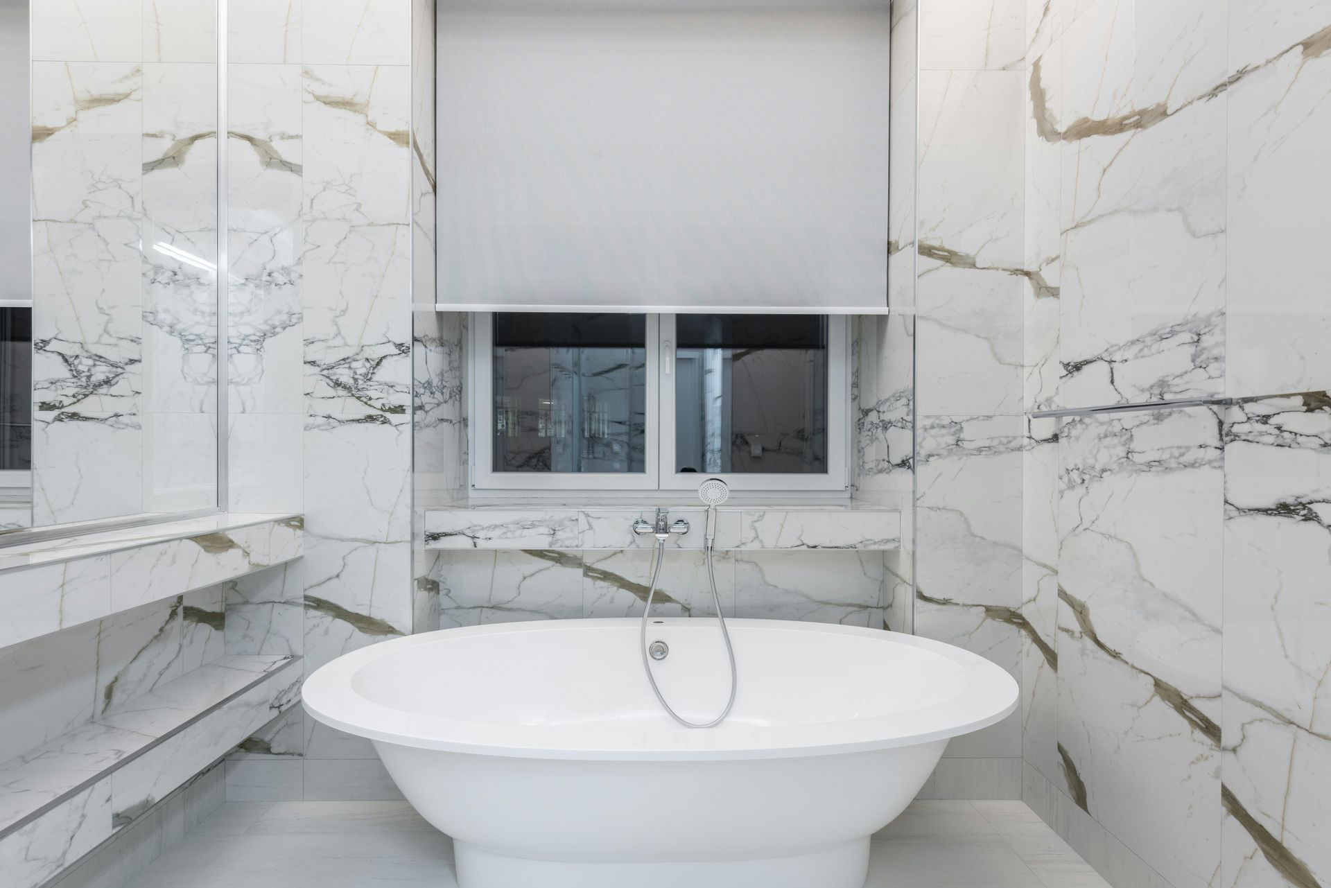 A modern white freestanding bathtub centered in a bathroom featuring white marble walls with gold veining and a window.
