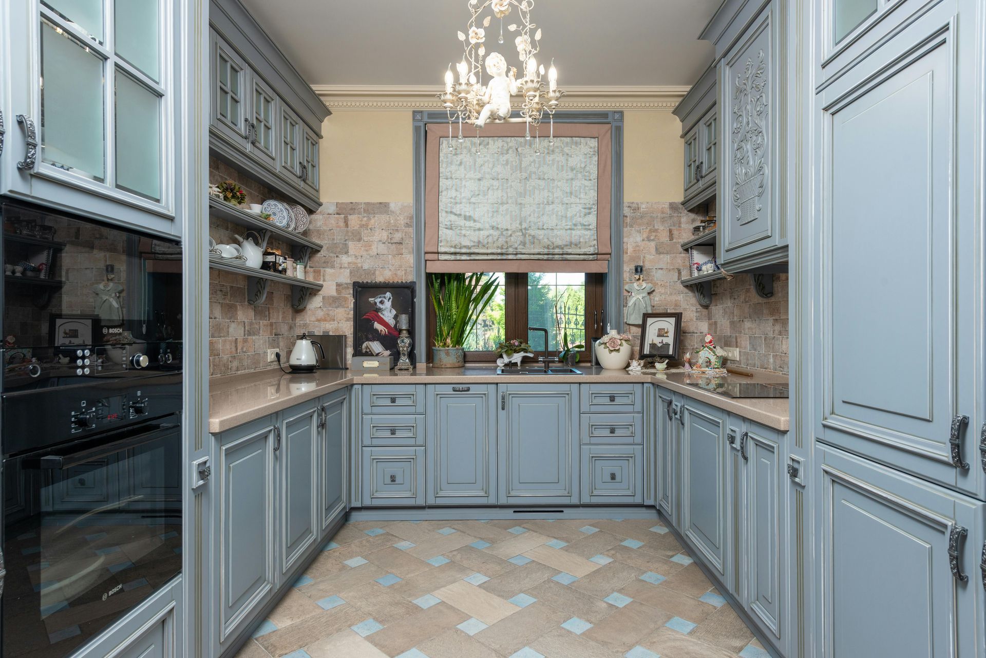 A U-shaped kitchen with dusty blue cabinets, a stone tile backsplash, patterned floor tiles, and a central chandelier.