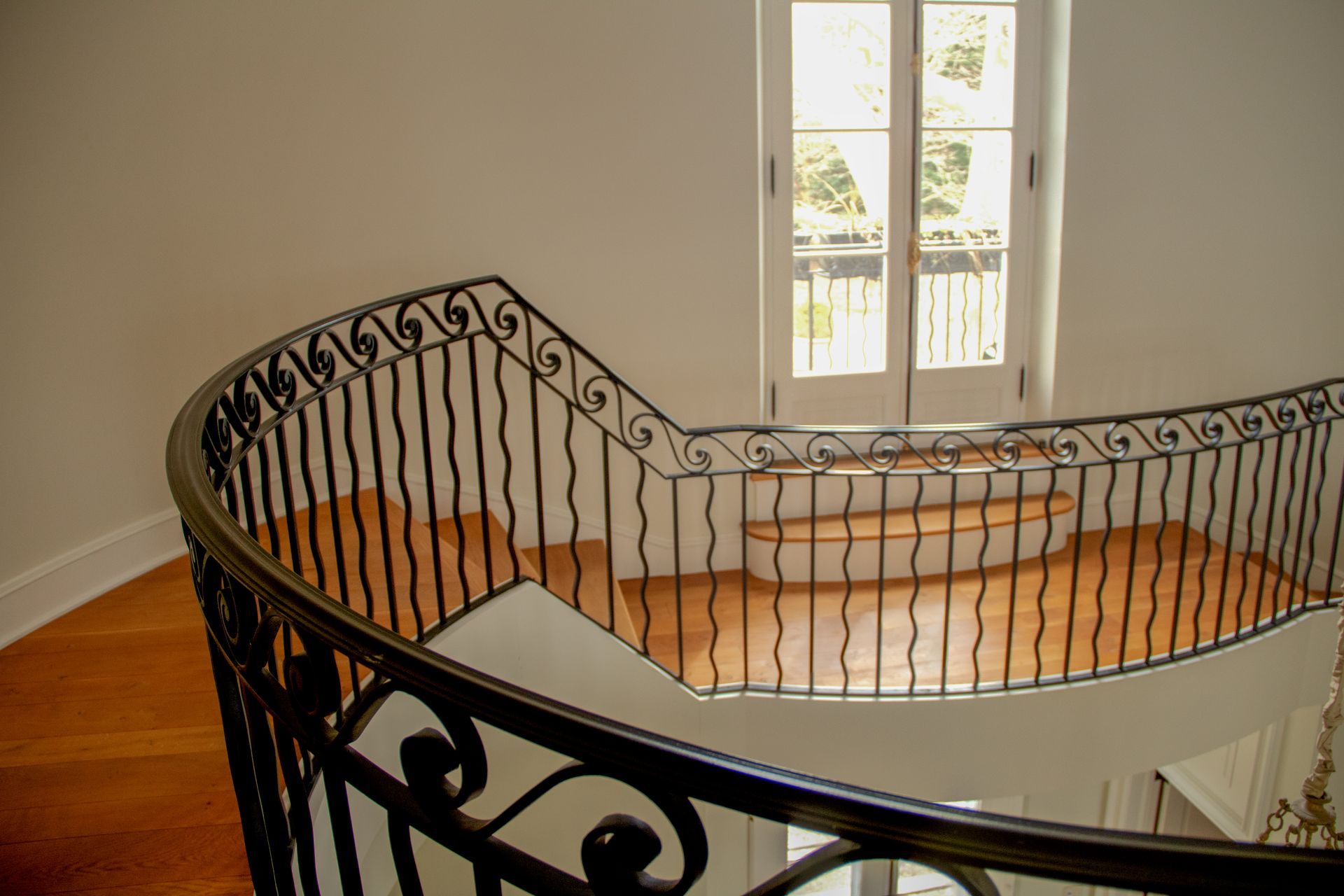 Curved staircase with black wrought iron railing, leading to a door with sunlight.