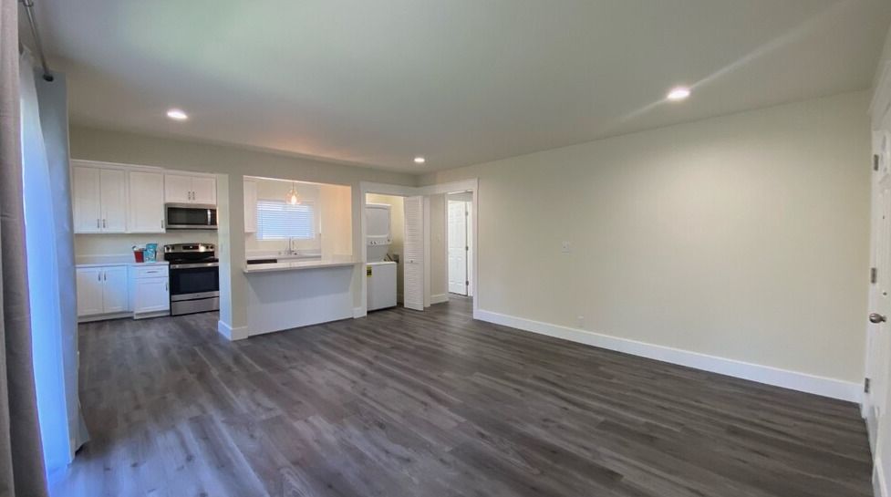 A living room with hardwood floors and a kitchen in the background.