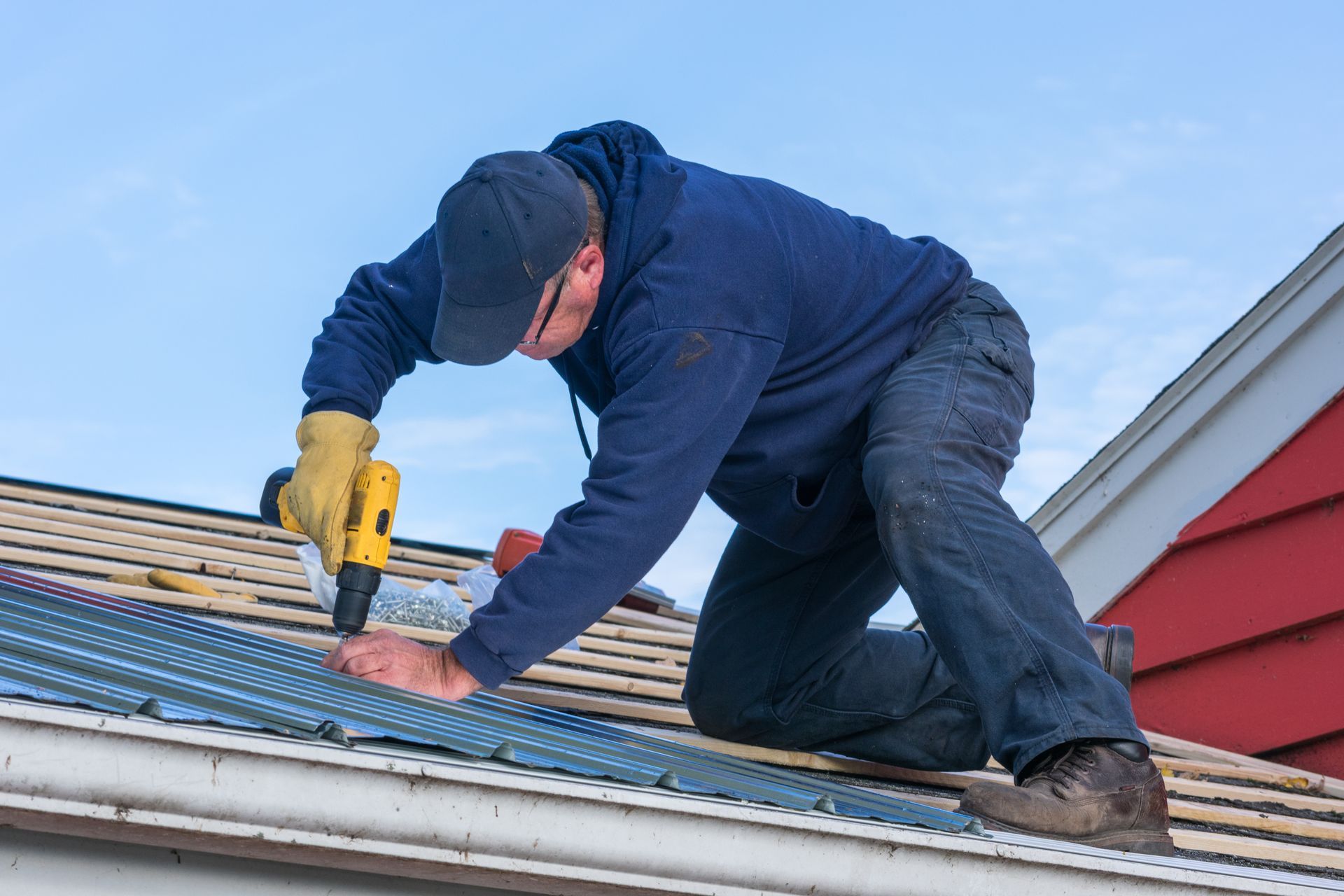 A professional roofer using a power drill to perform a roof leak repair on a metal rooftop. A professional roofer using a power drill to perform a roof leak repair on a metal rooftop.