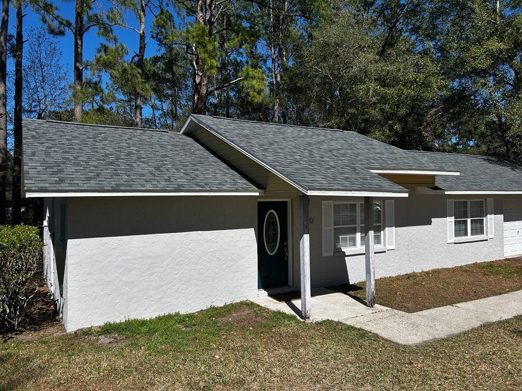 A light gray house with a dark gray roof sits on a grassy area surrounded by trees.