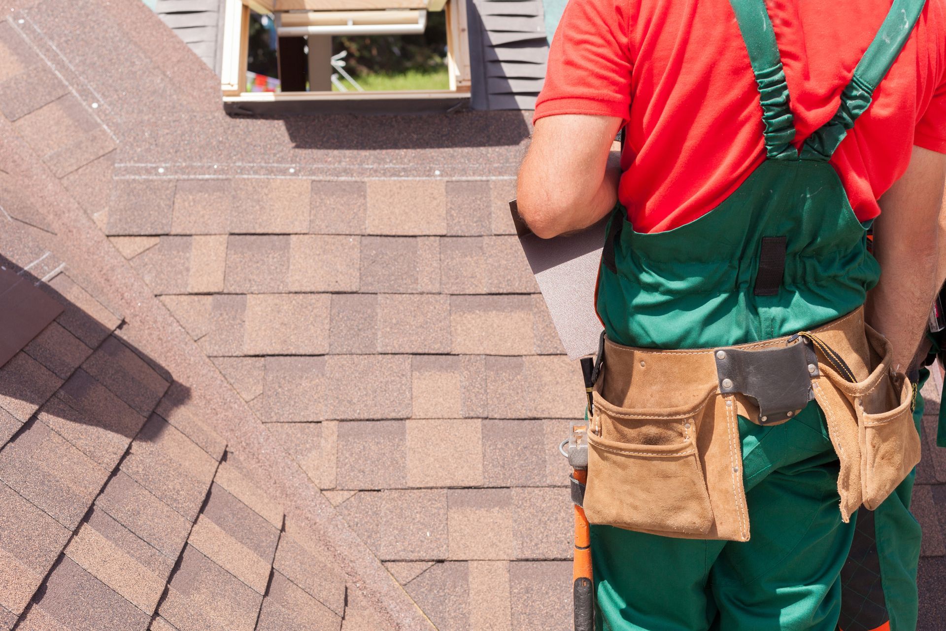Roofer in red shirt and green overalls repairing brown shingle roof near open skylight window.