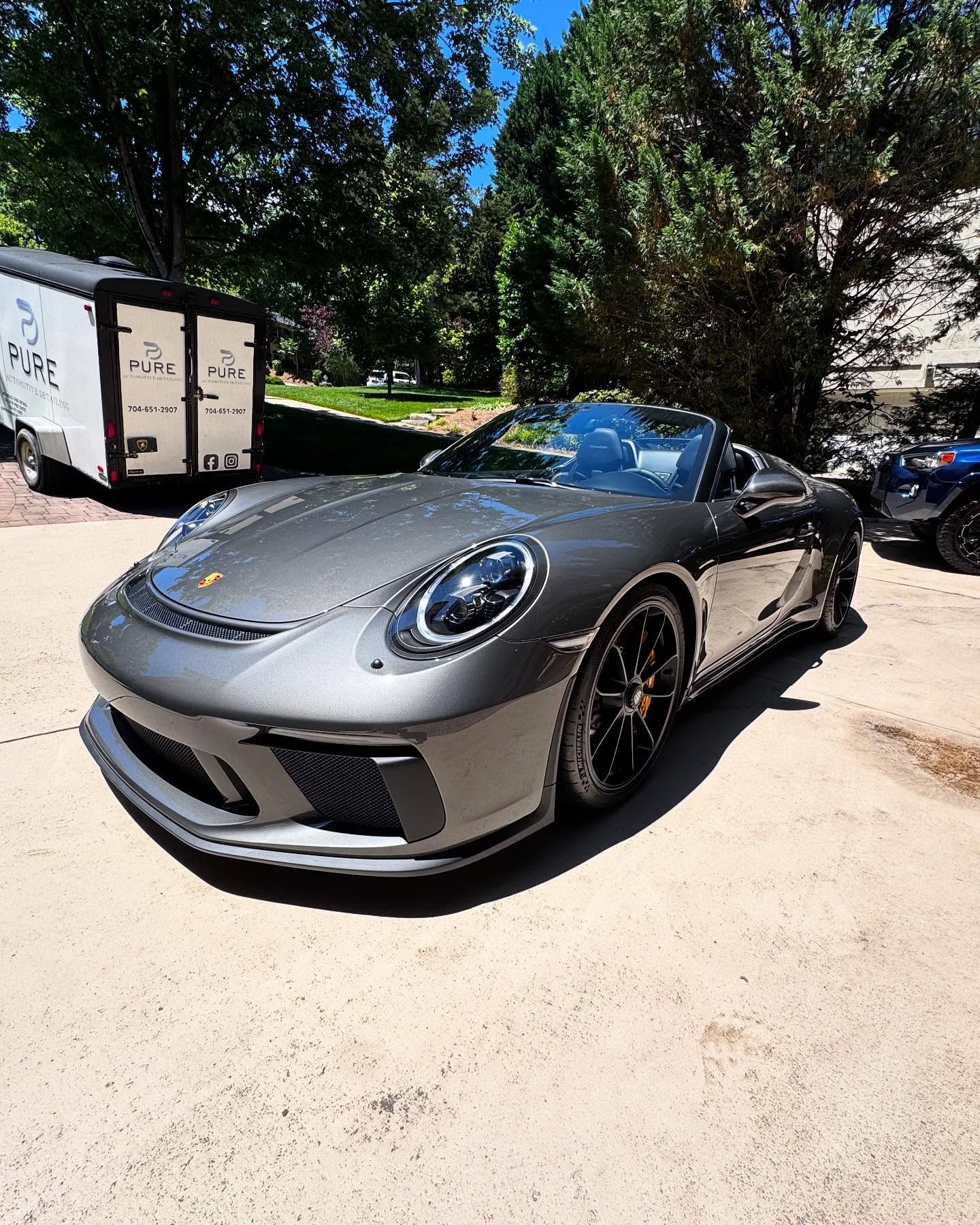 A gray porsche 911 convertible is parked in a driveway next to a trailer.