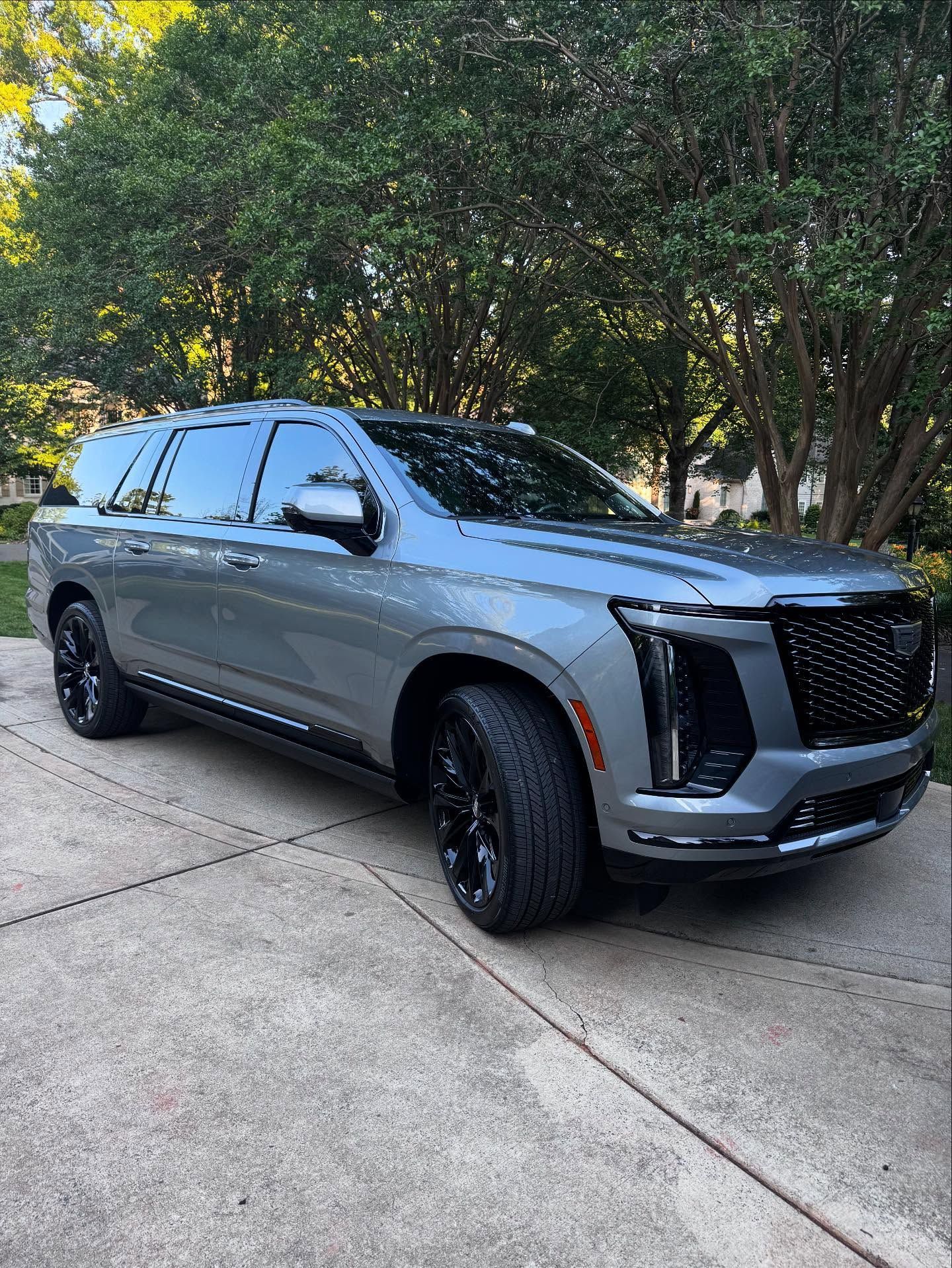 A gray cadillac escalade is parked in a driveway next to trees.