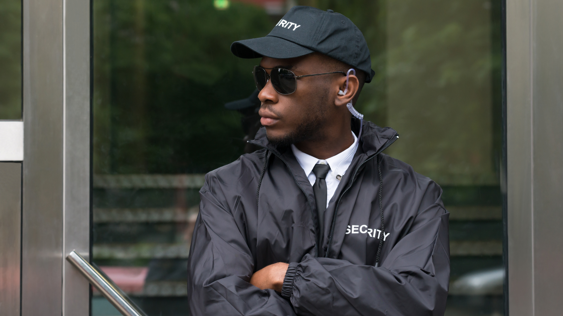 A security guard wearing a hat and sunglasses is standing in front of a glass door.