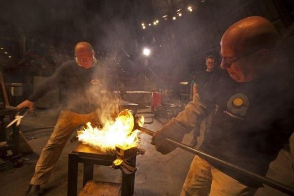 Two glassblowers working with molten glass, flames, dark setting.
