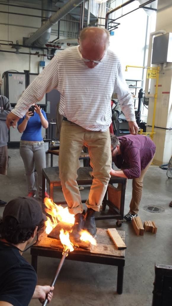 Glass Artist, John de Wit, stands on a burning platform. Two people watch/assist in a workshop.