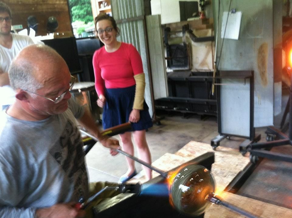 Glass Artist, John de Wit, blowing glass sphere with two onlookers in workshop.