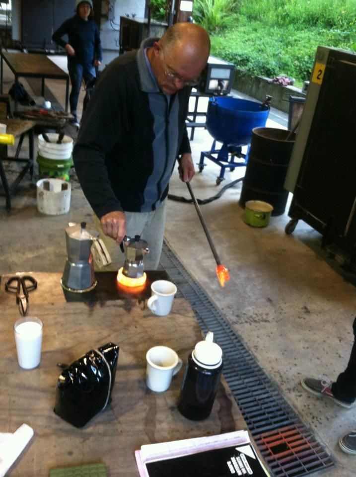 Glass Artist, John de Wit, heats glass rod over a small flame, while making coffee on a workbench with various objects.