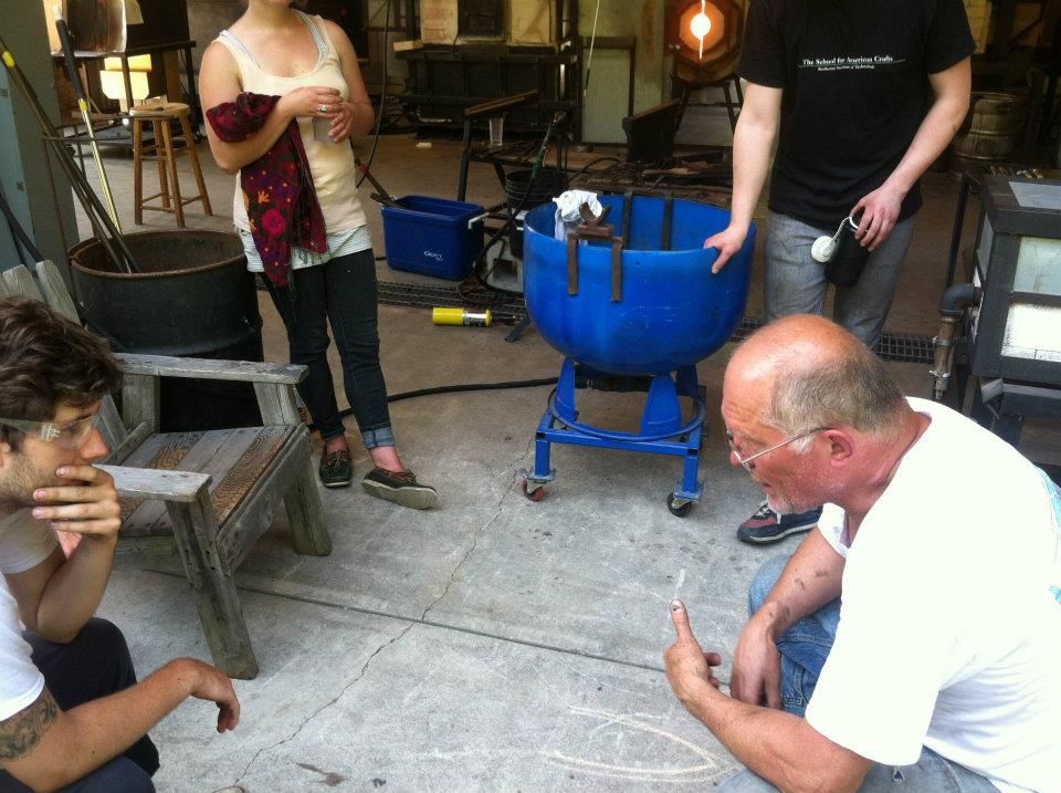 People in a glass-blowing workshop; Glass Artist, John de Wit, points at the floor. A blue container on wheels sits center.