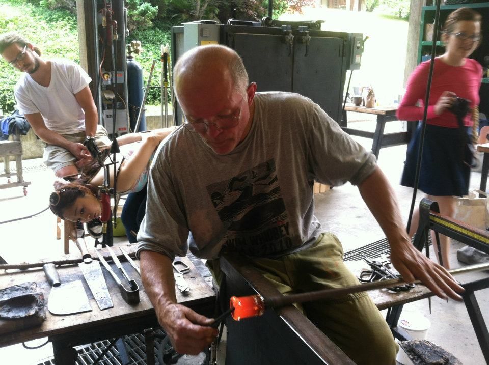 Glass Artist, John de Wit, working with molten glass, others in a glassblowing studio.